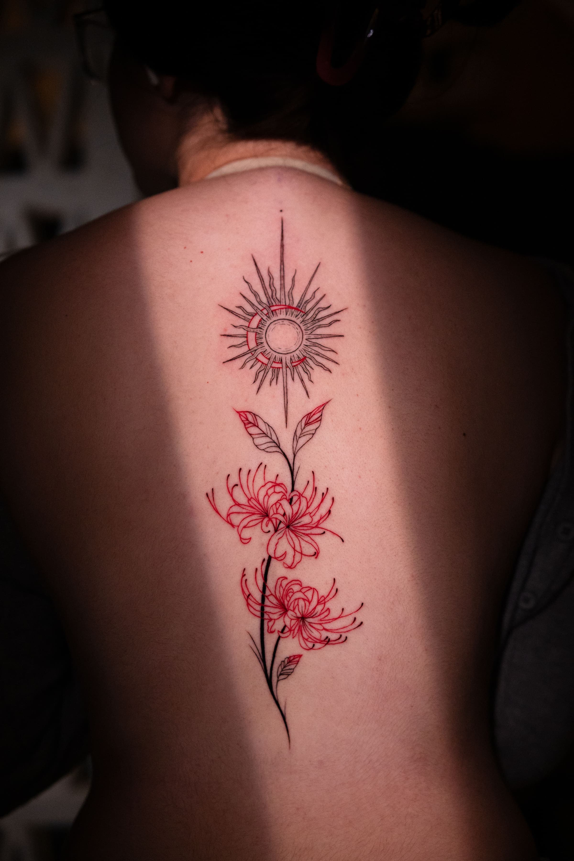 Fine line tattoo of red spider lilies and a geometric sun along the spine, by Rivas Indeed, tattoo artist in San Juan, Puerto Rico